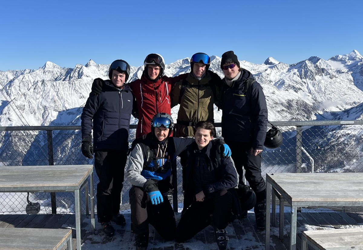 Gruppe in Skiausrüstung auf einer Aussichtsplattform vor verschneiten Bergen